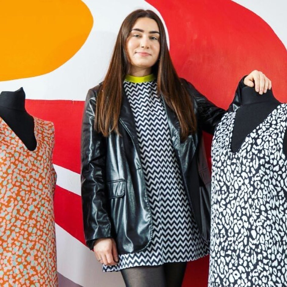 Young woman with long dark brown hair is standing with two mannequins dressed in dresses she made.