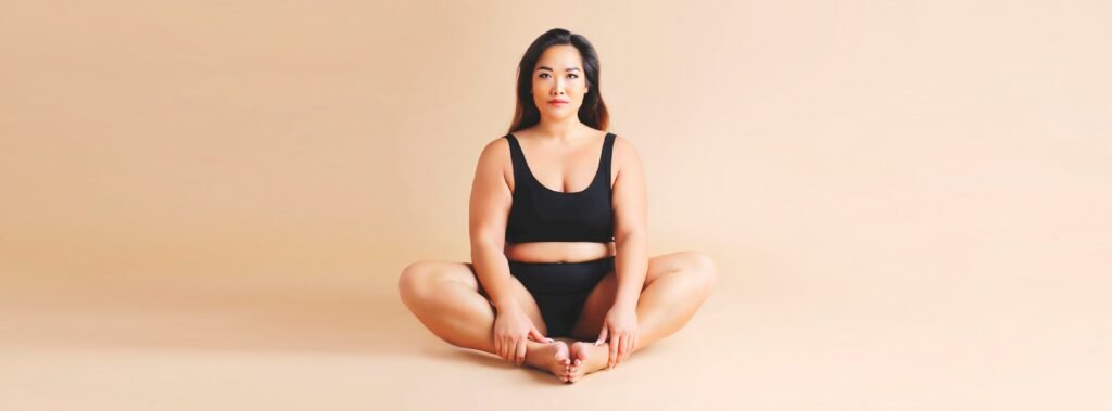 Young woman sitting in her black underwear with feet touching and legs out to the side. She is wearing her Amala period pants.