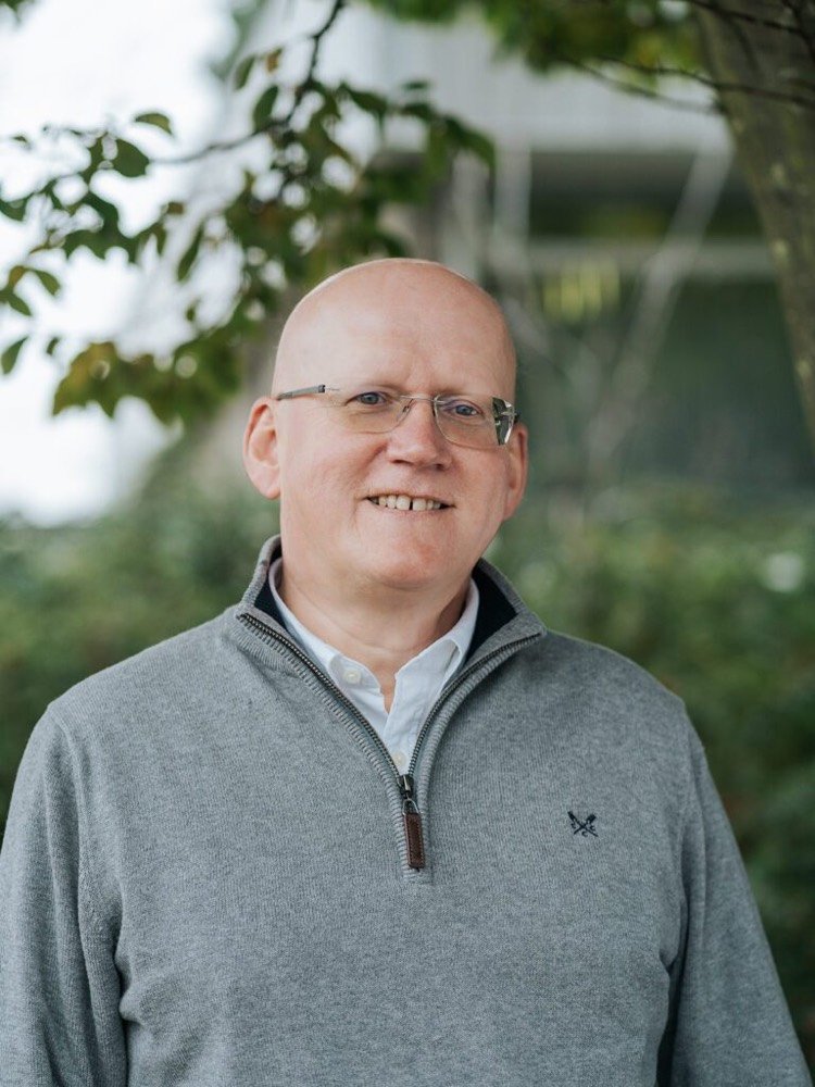 Headshot of Donald McPherson. Donald wears glasses and a grey zip-up jumper. He stands in front of some greenery.