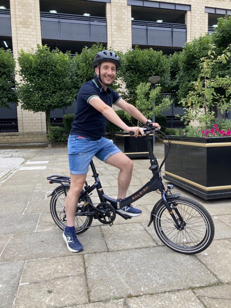 Matt (a man wearing a black polo shirt and jean shorts) sits on an electric bike