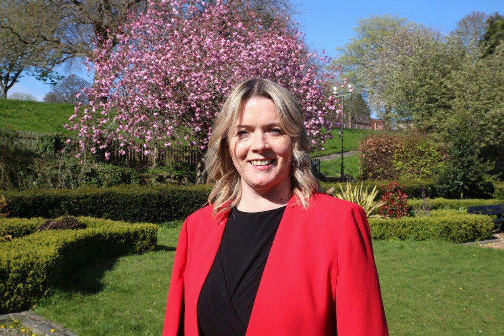 A photo of Kirsty Thomson. In the photo, Kirsty is wearing a black dress with a red blazer. She is standing in the garden outside of Dudhope Castle and is in front of a blossoming tree