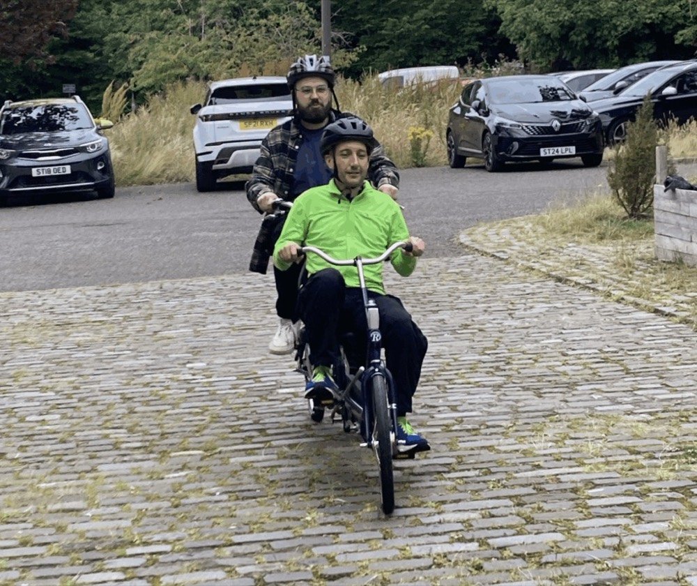 Two men are riding a tandem bike on a cobbled patch of road
