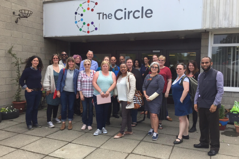 A group of 21 people standing together in front of a building underneath a sign with The Circle logo on it