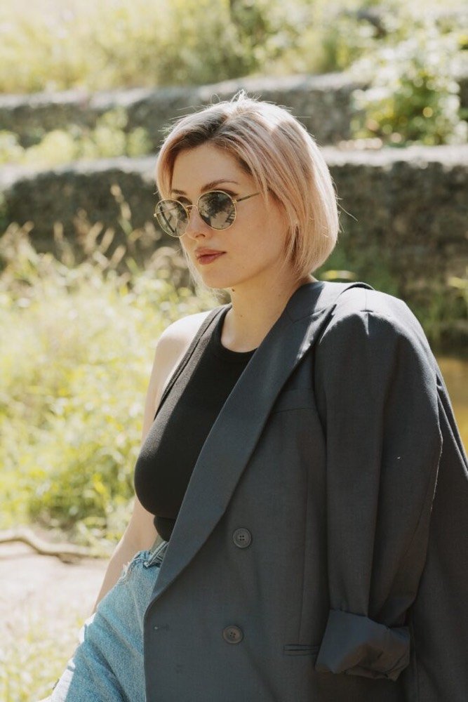 Yulia SKULSKIKH in a garden. The photo is taken side on with Yulia looking away from the camera. She is wearing a charcoal suit jacket on one shoulder, a black sleeveless top and jeans. She has one hand in her pocket and is wearing round sunglasses. She has chin length straight blonde hair.