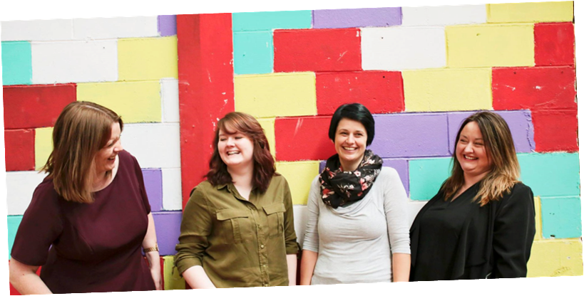 The ACK team - Kirsty, Jenny, Clare and Fiona - standing together in front of a multi-coloured brick wall and laughing